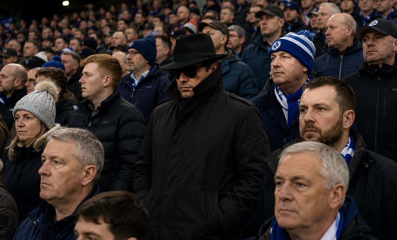 A crowd at a sports stadium with a man in a black coat, hat, and sunglasses standing among fans in winter jackets.